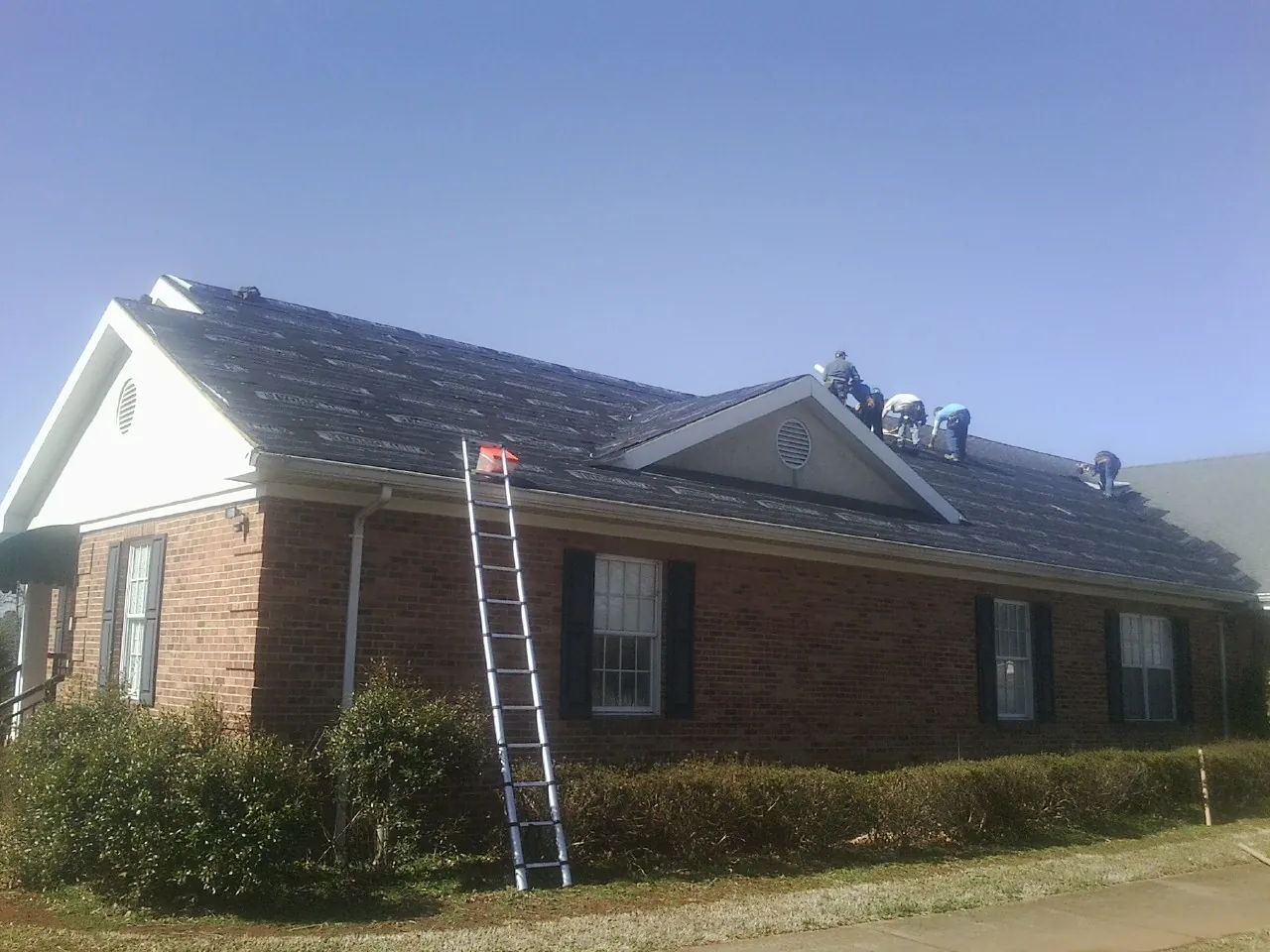 Crew tearing shingles off a brick ranch roof with a ladder set — Emergency Roof Repair work by our crew in Table Grove