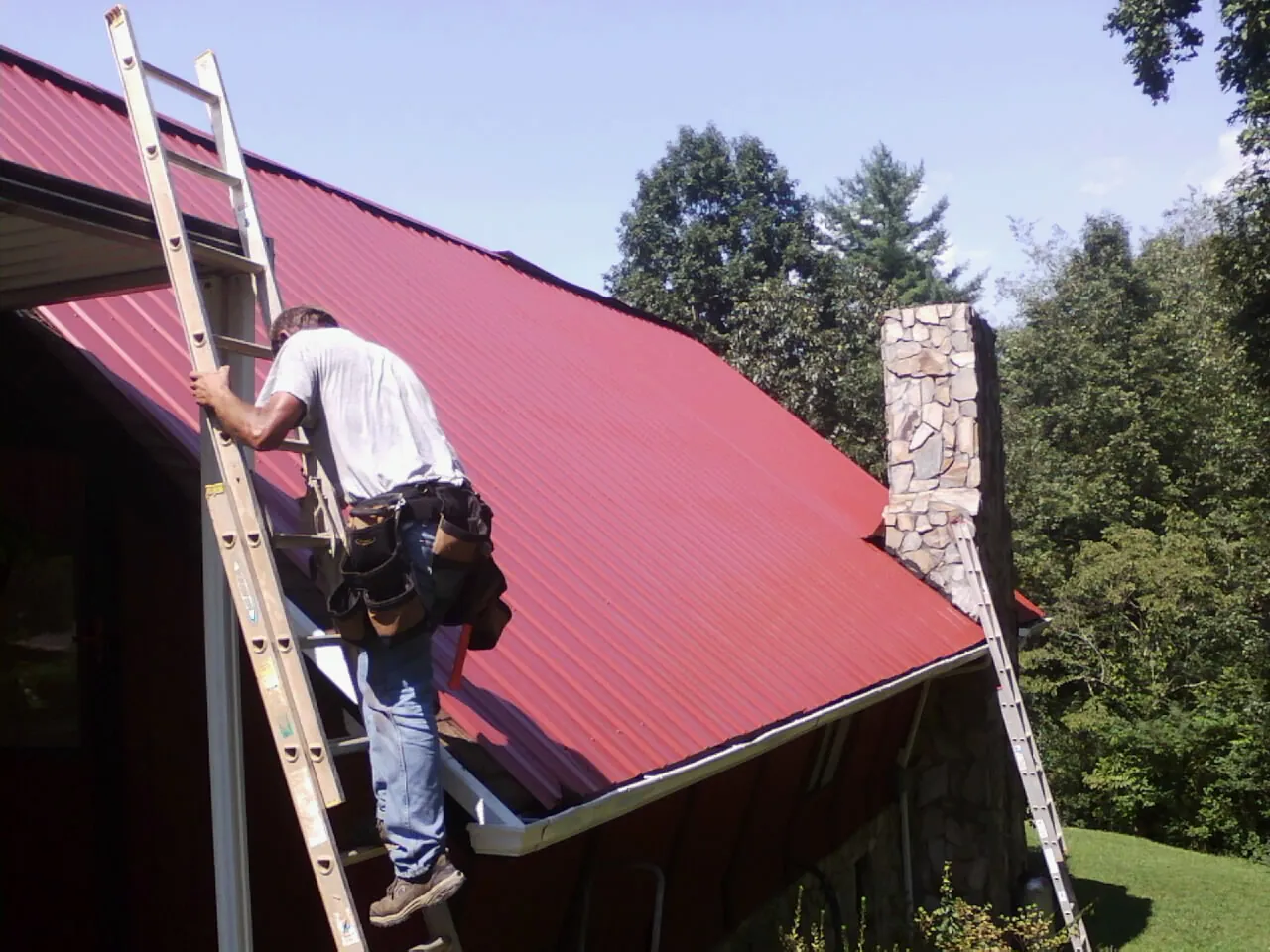 Roofer on a ladder beside a red metal roof and stone chimney — Soffit & Fascia Repair by Roofing Brothers in Table Grove
